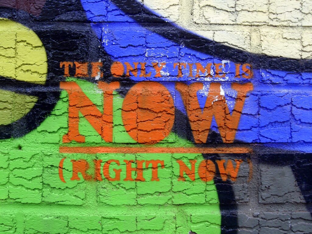 Bunt besprühte Mauer, darauf Text: The only time is now (right now)