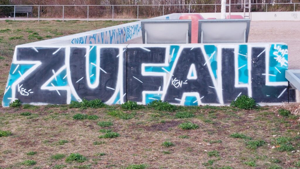 Mauerstück auf einer Wiese, auf dem das Wort ZUFALL gesprayt ist